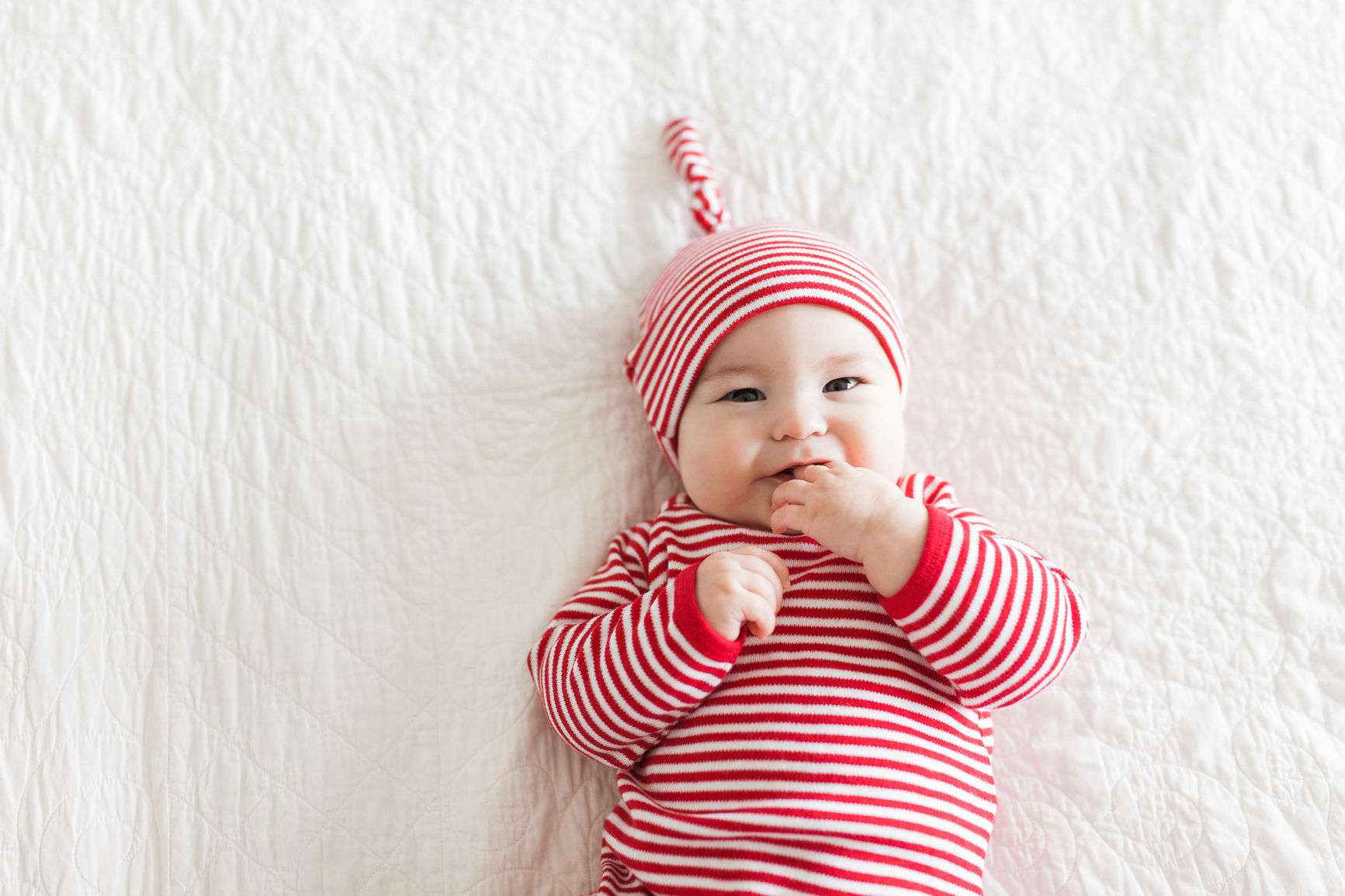 Liegendes Baby in rot-weiß-gestreiftem Body und Mütze knabbert an seiner Hand.