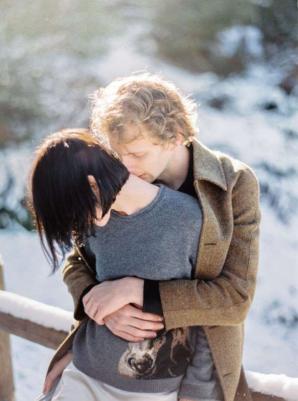 Mann umarmt Frau in Winterlandschaft von hinten und küsst ihren Nacken.