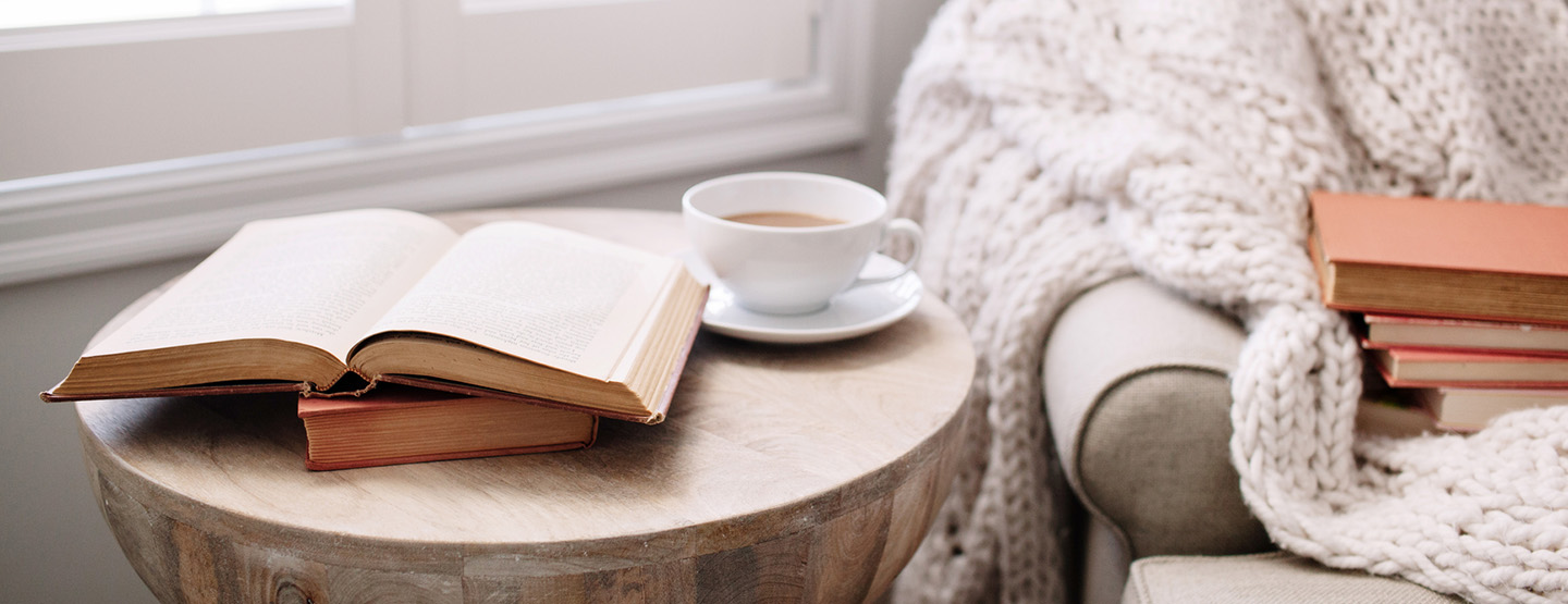 Holztisch mit Büchern und Tasse Kaffee, neben gemütlichem Sessel laden ein zum Cocooning