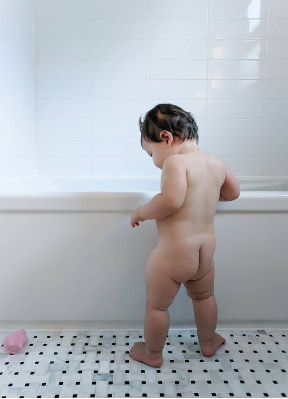 ein Kleinkind das nackt vor der Badewanne steht und ins Wasser schaut