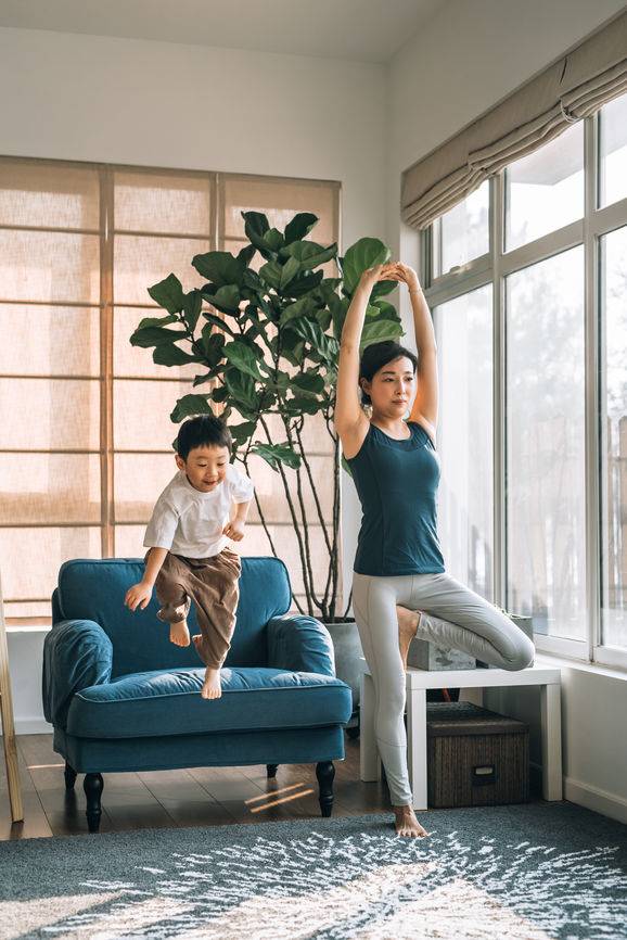 Eine Mutter die auf einer Turnmatte im Wohnzimmer eine Yoga-Übung praktiziert und ihr Sohn sitzt auf ihrem Rücken und lacht