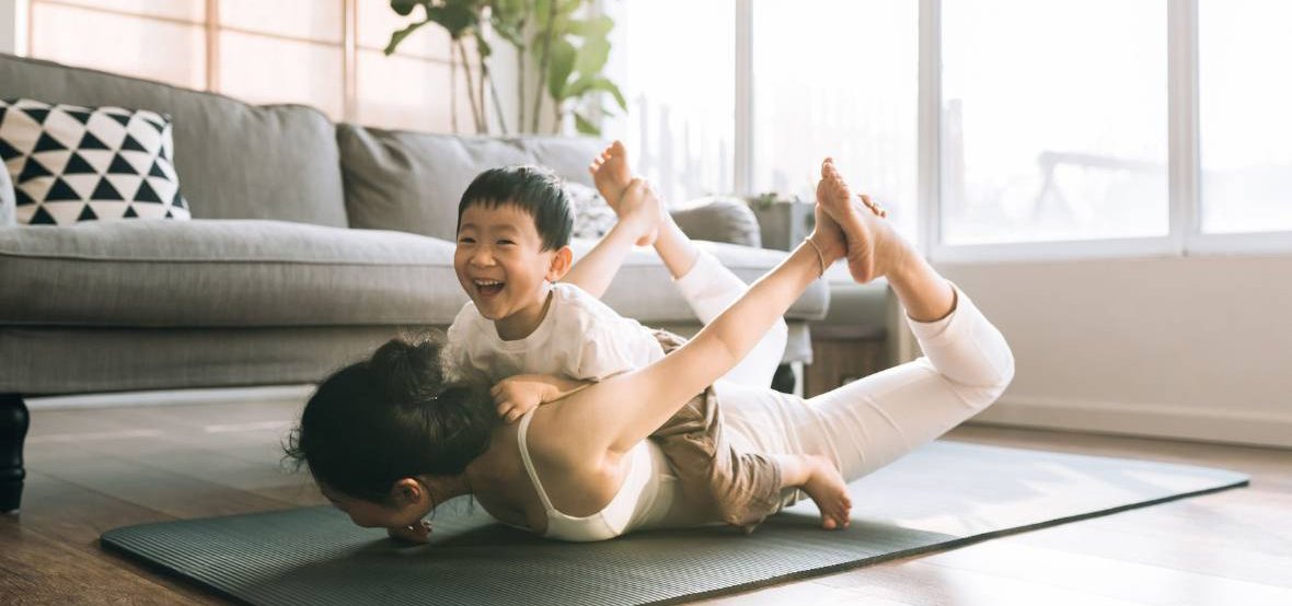 Eine Mutter die auf einer Turnmatte im Wohnzimmer eine Yoga-Übung praktiziert und ihr Sohn sitzt auf ihrem Rücken und lacht