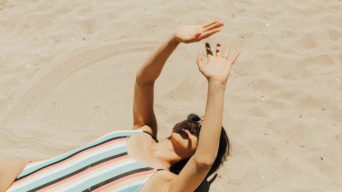 Junge Frau liegt im Badeanzug am Strand und hält ihre Arme in die Höhe, um ihr Gesicht vor der Sonne zu schützen