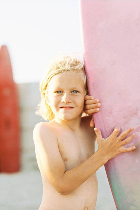 Ein kleiner Junge in Badeshorts der ein Surfbrett in den Händen hält