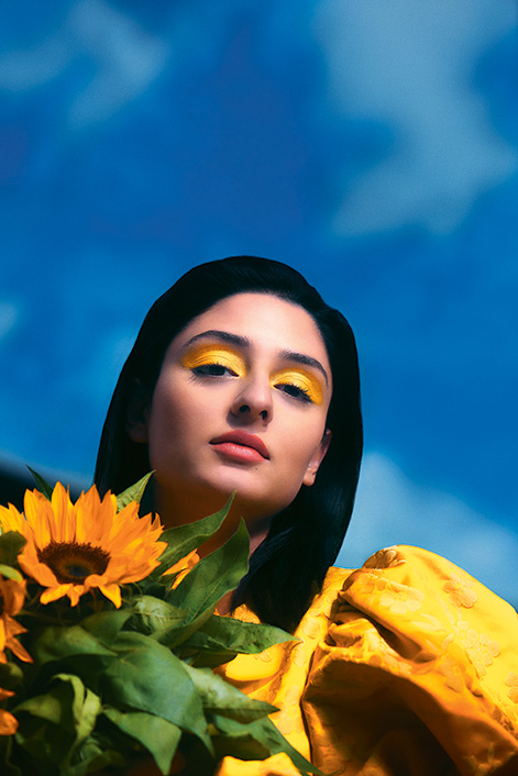 Frau in Gelb gekleidet mit Sonnenblumen und dunklem Haar vor strahlend blauem Himmel