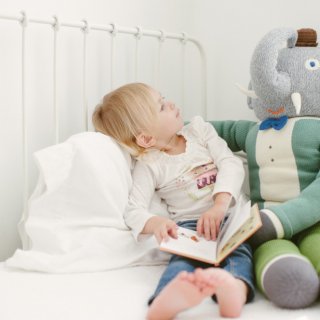 Mädchen mit Buch über Kinderfragen schaut fragend zum großen Stoffelefant neben ihr.