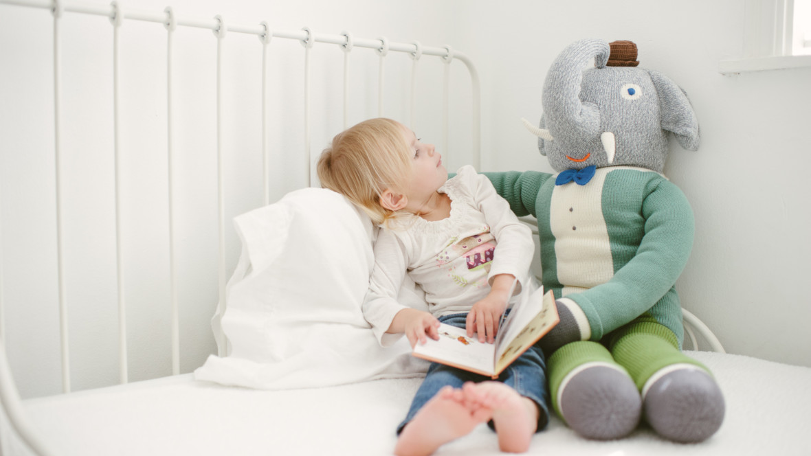 Mädchen mit Buch über Kinderfragen schaut fragend zum großen Stoffelefant neben ihr.