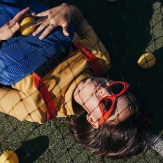 Junge Frau mit roter Sonnenbrille und blauem Kleid liegt hinter Tennisnetz bei Sportspielen am Boden.