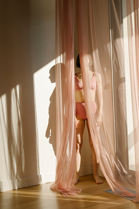Frau in rosa Unterwäsche steht in der Ecke einer Wohnung hinter einem durchsichtigen rosa Vorhang