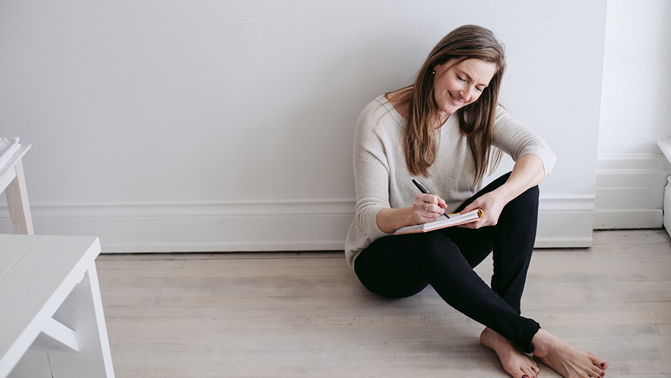 Langhaarige Frau in den Wechseljahren schreibt am Boden sitzend in ein Buch