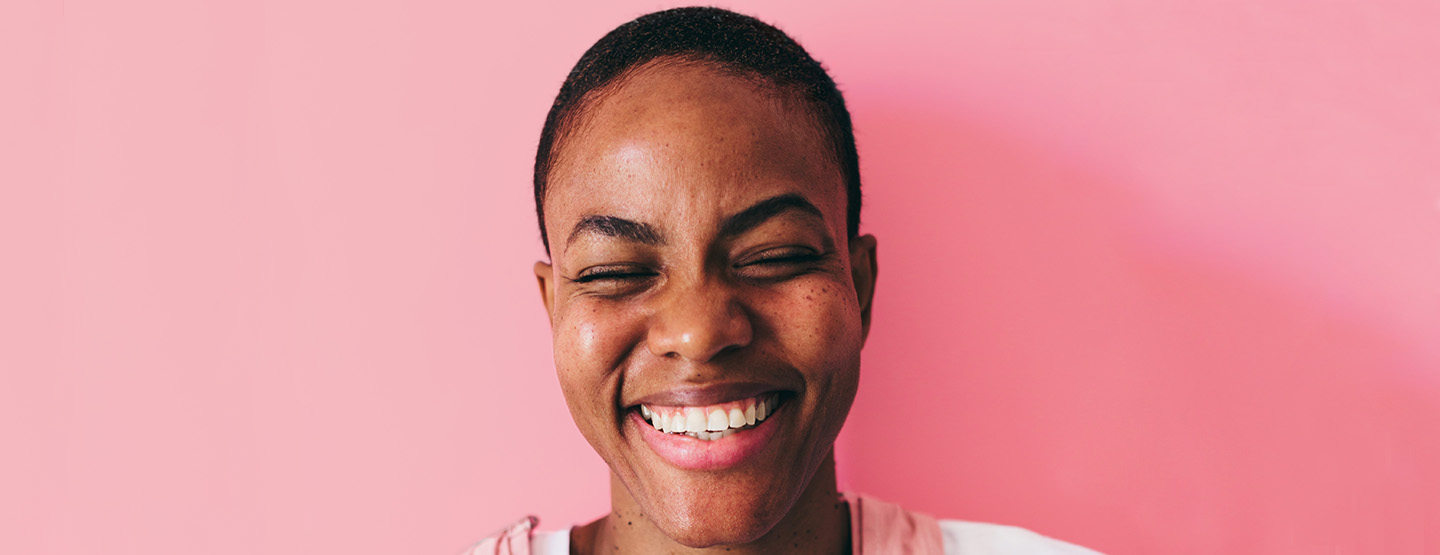 Humor lernen: Frau mit Buzz Cut vor rosa Hintergrund, die mit geschlossenen Augen herzlich lacht