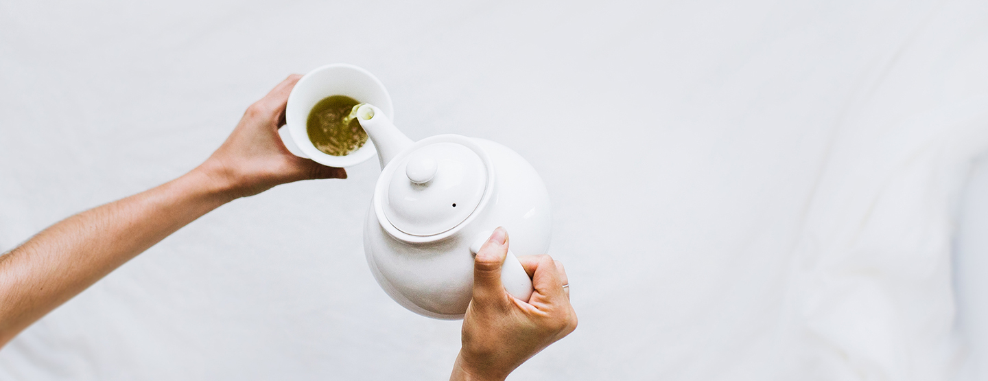 Jemand schenkt grünen Tee aus einer weißen Kanne in eine Tasse