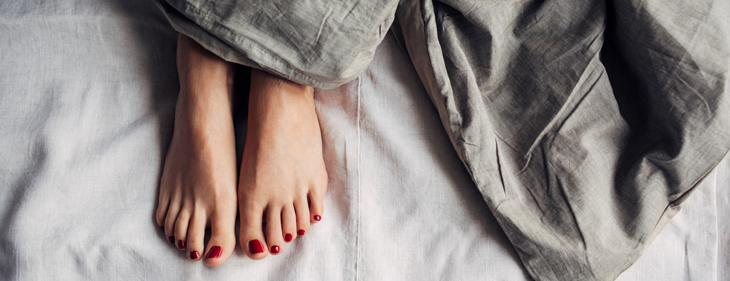 Füße mit rot lackierten Nägeln lugen unter grauer Bettdecke hervor