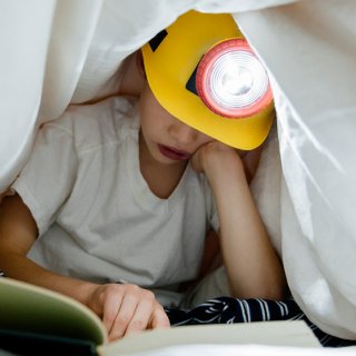 Lesendes Kind unter Bettdecke mit Bauarbeiterhelm und Stirnlampe