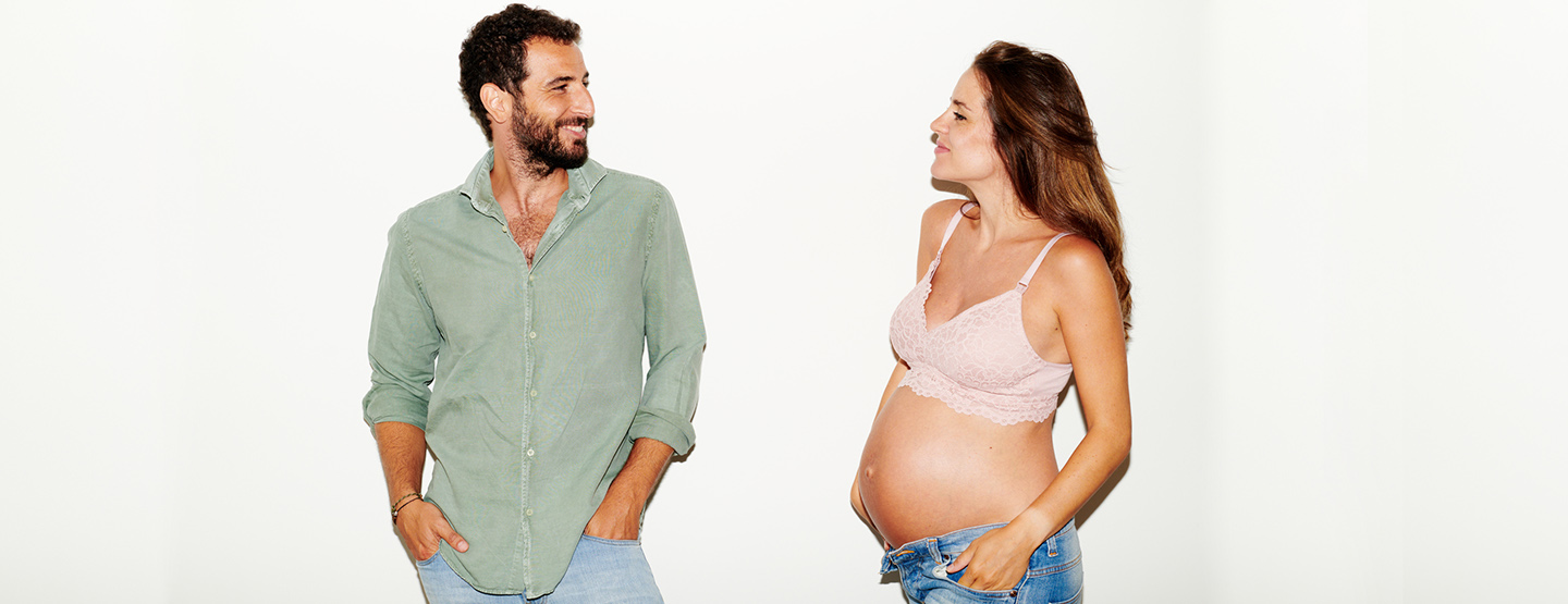 Schwangere mit nacktem Bauch steht neben ihrem Partner und lächelt ihn an