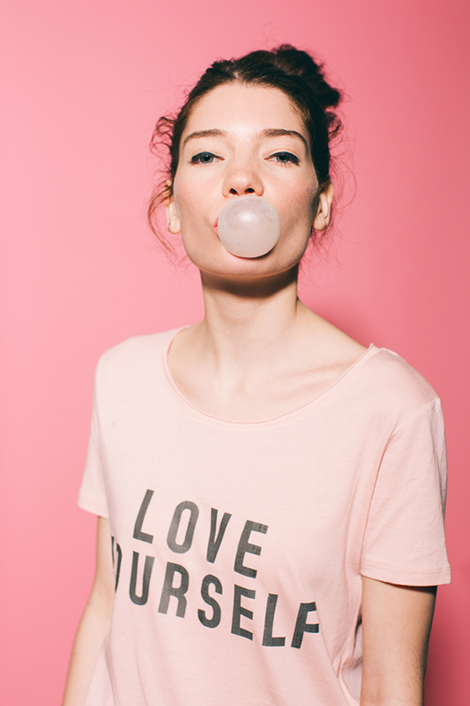 Frau in rosa T-Shirt mit Aufdruck "Love Yourself" macht Kaugummi-Blase