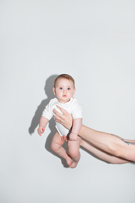 Baby in weißem Body wird von Armen vor weißem Hintergrund gehalten