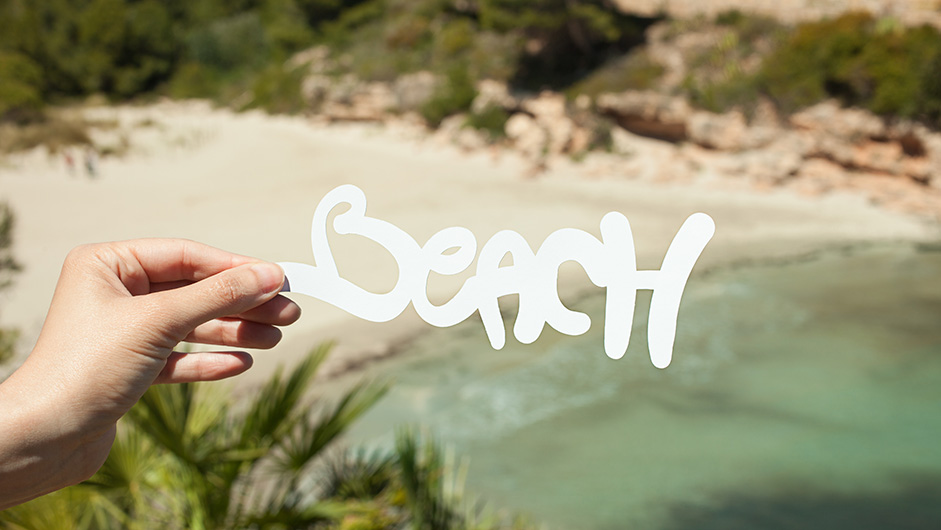 Hand hält Schriftzug "Beach" über den Hintergrund mit Strand und Meer