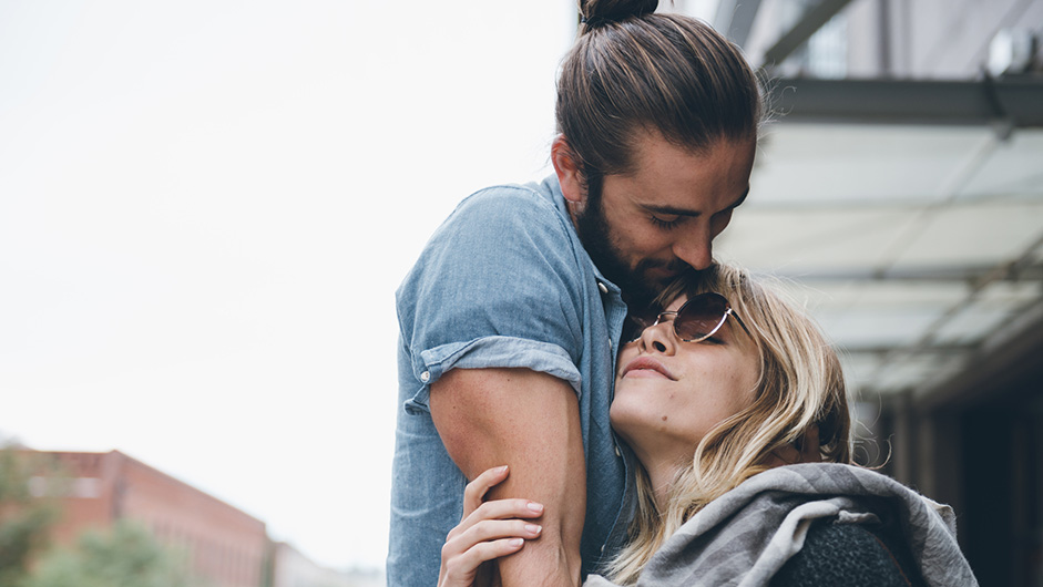 Mann mit zusammengebundenem, langem Haar und Frau mit Sonnenbrille liebkosen sich